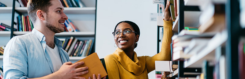 Male and female entrepreneurs standing next to one another by two sets of bookshelves. Discussing ways to use their business plan.